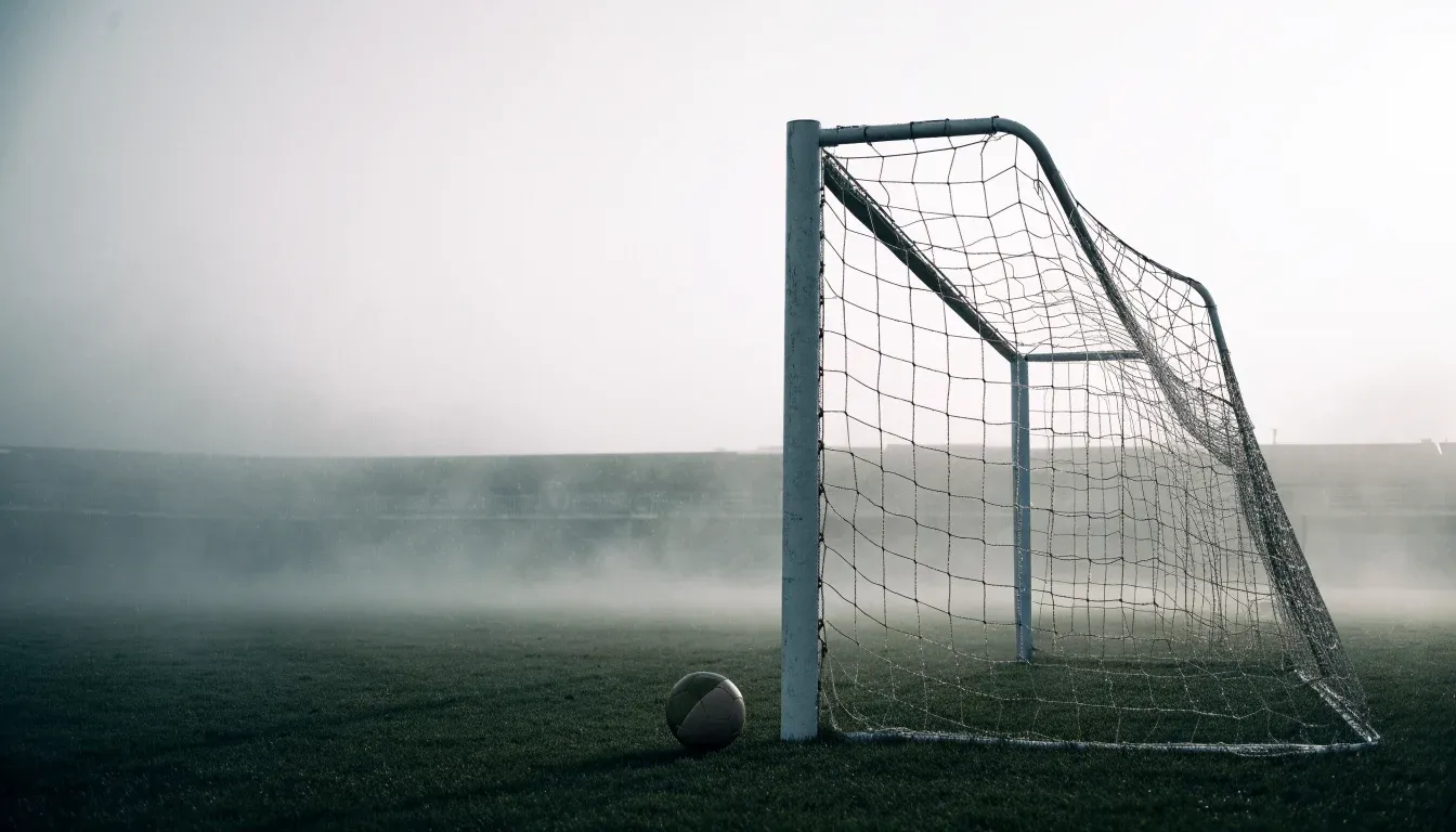Ein Fußballtor im Nebel eines frühen Morgens, das Netz hängt feucht, auf dem Rasen davor liegt ein einzelner Ball.